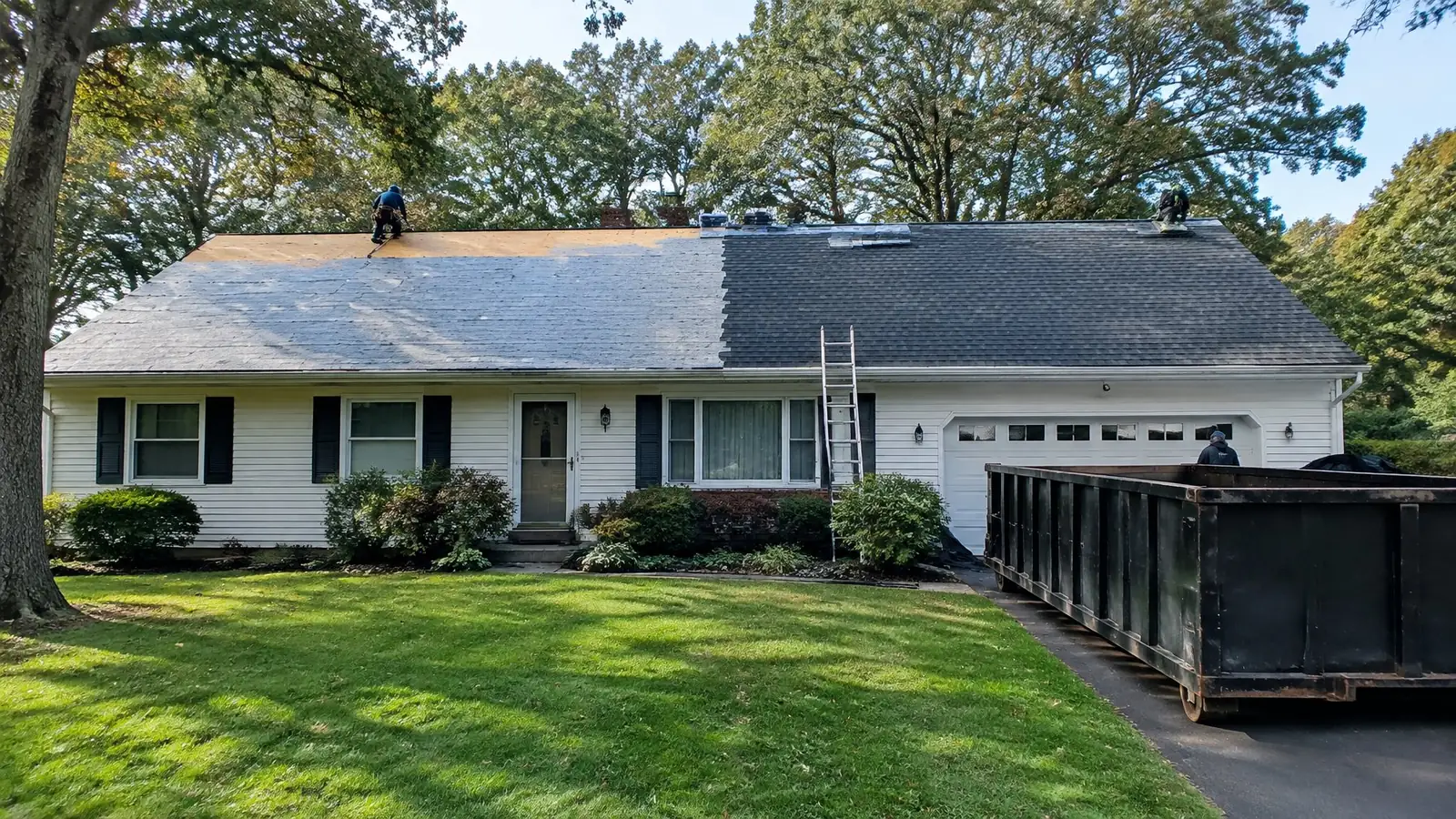 Ranch house mid-replacement: one half of the roof in new dark shingles, the other half still old, with a dumpster in the driveway